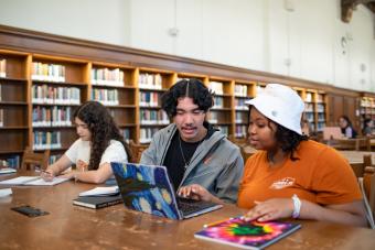 Three students sitting at a large desk wokring on a laptop.